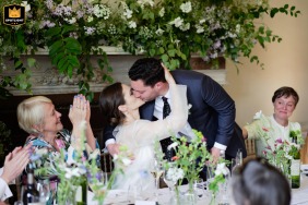At Gunnersbury Park in London, a bride kisses the standing groom after his speech, as mothers applaud. The scene includes head table florals in the foreground and overhead foliage and florals in the background.